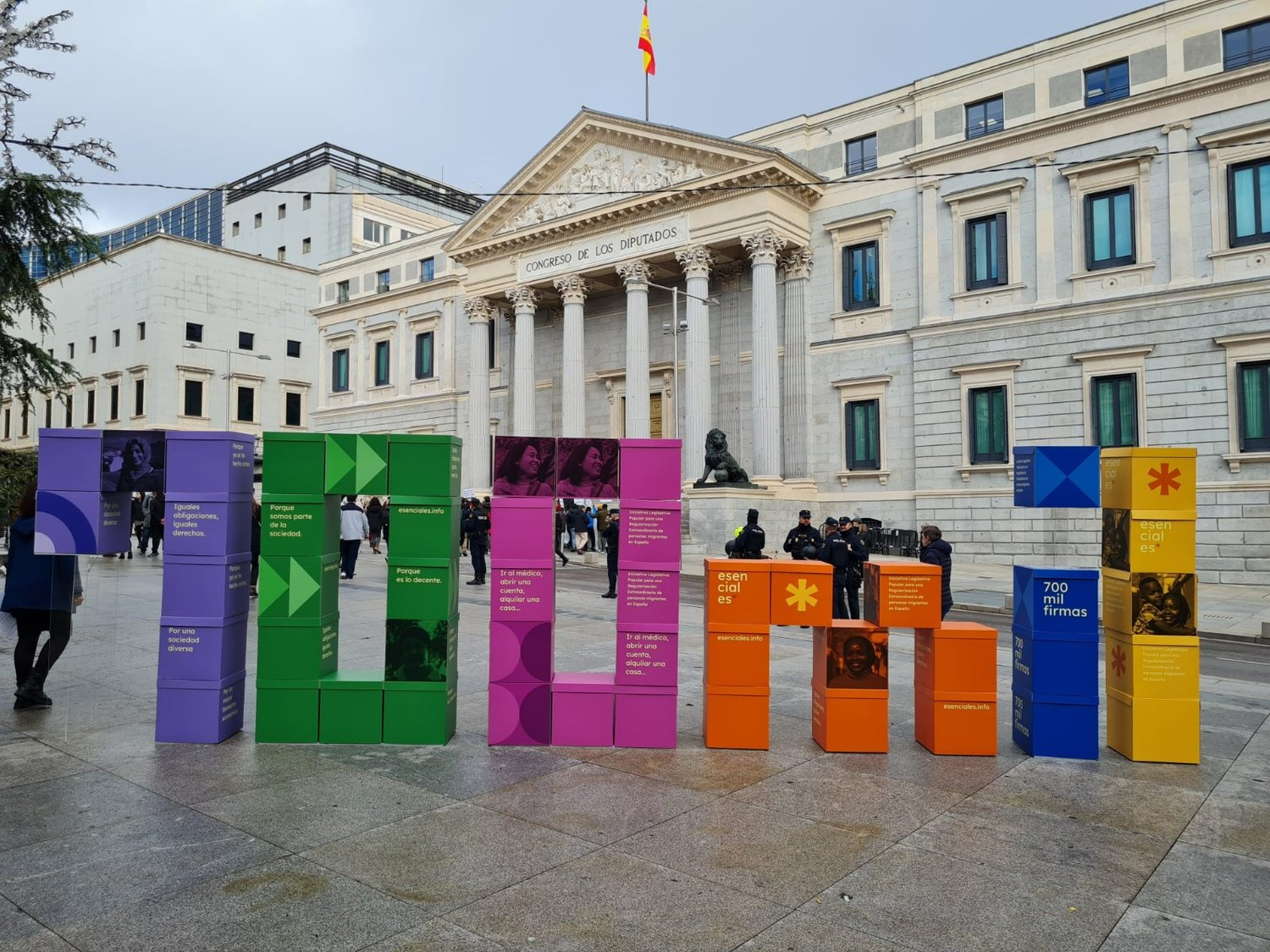 Las letras ILP formadas con grandes cajas de colores de la campaña Esenciales frente a la fachada del Congreso de los Diputados