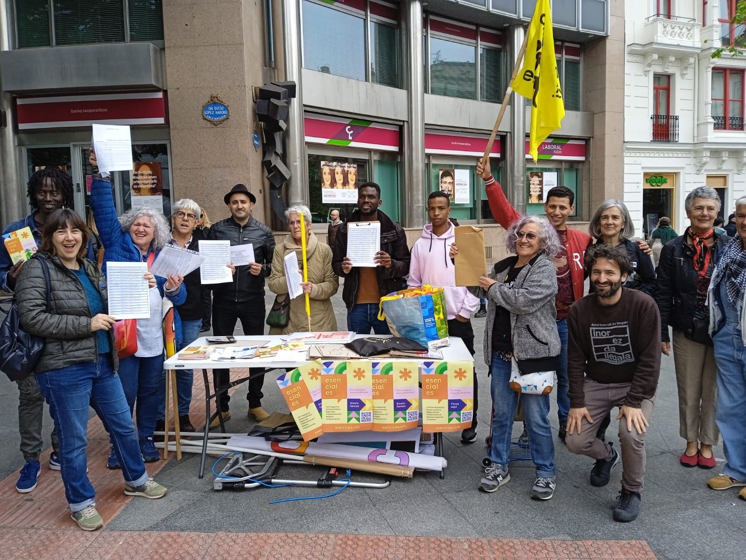 Un grupo de militantes posa en la calle mostrando pliegos de firmas junto a una mesa con carteles de la campaña Esenciales en Bilbao